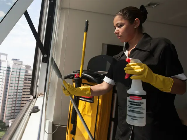 Faxineira de uniforme e luvas amarelas limpando a janela de um apartamento com carrinho de limpeza