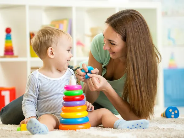 Babá pajem sorrindo para um bebê que brinca com argolas coloridas no tapete do quarto